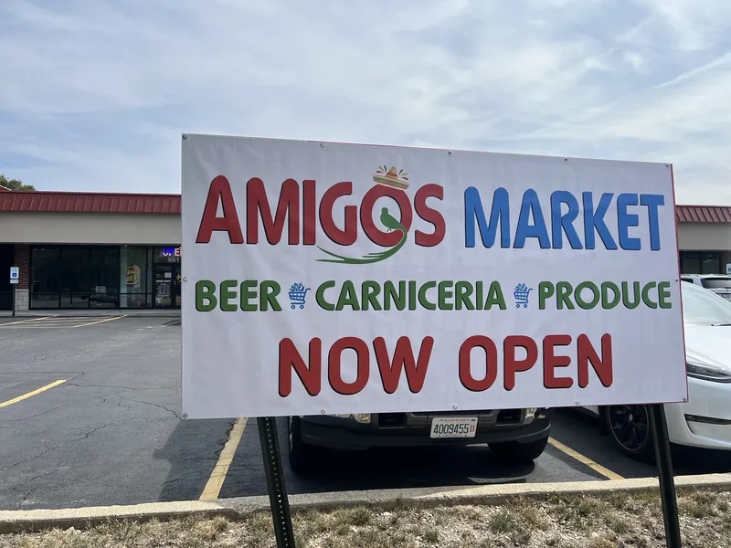 Large white banner reading Amigos Market, Beer, Carniceria, Produce, Now Open, mounted in a parking lot