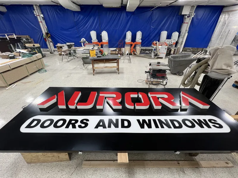 Large black sign panel reading Aurora Doors and Windows in red and white lettering laid out in a workshop