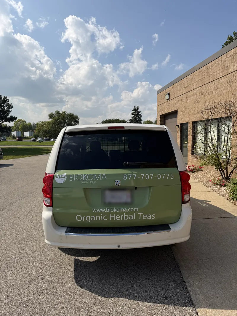 Rear of a white minivan with a green graphic panel showing the Biokoma logo, phone number and website