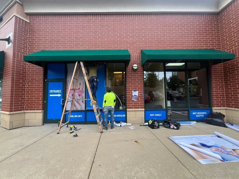 Installer on a ladder applying printed dental graphics to storefront windows under green awnings