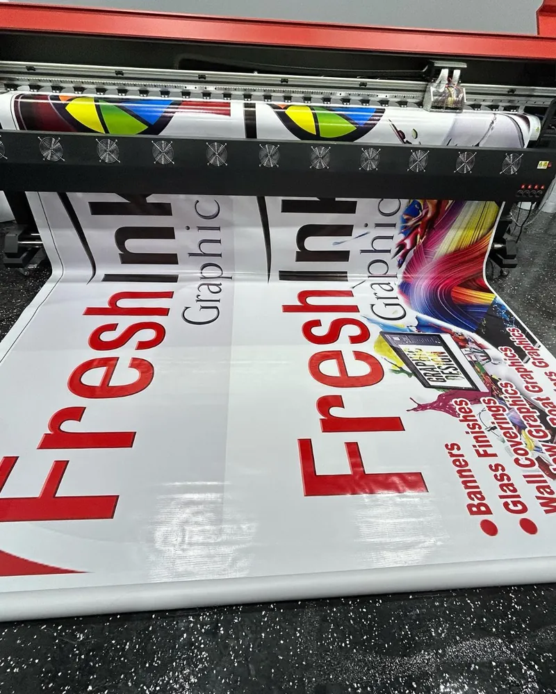 Wide-format printer printing a Fresh Ink Graphics banner listing banners, glass finishes and wall coverings