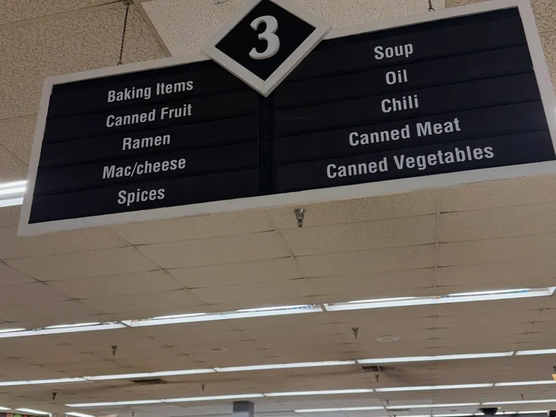 Black hanging aisle sign numbered 3, listing baking items, canned goods, soup, oil and spices