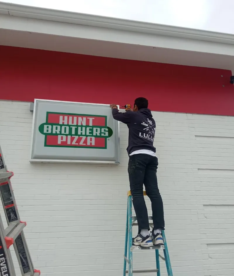 Installer on a ladder mounting a lit Hunt Brothers Pizza sign on a store wall