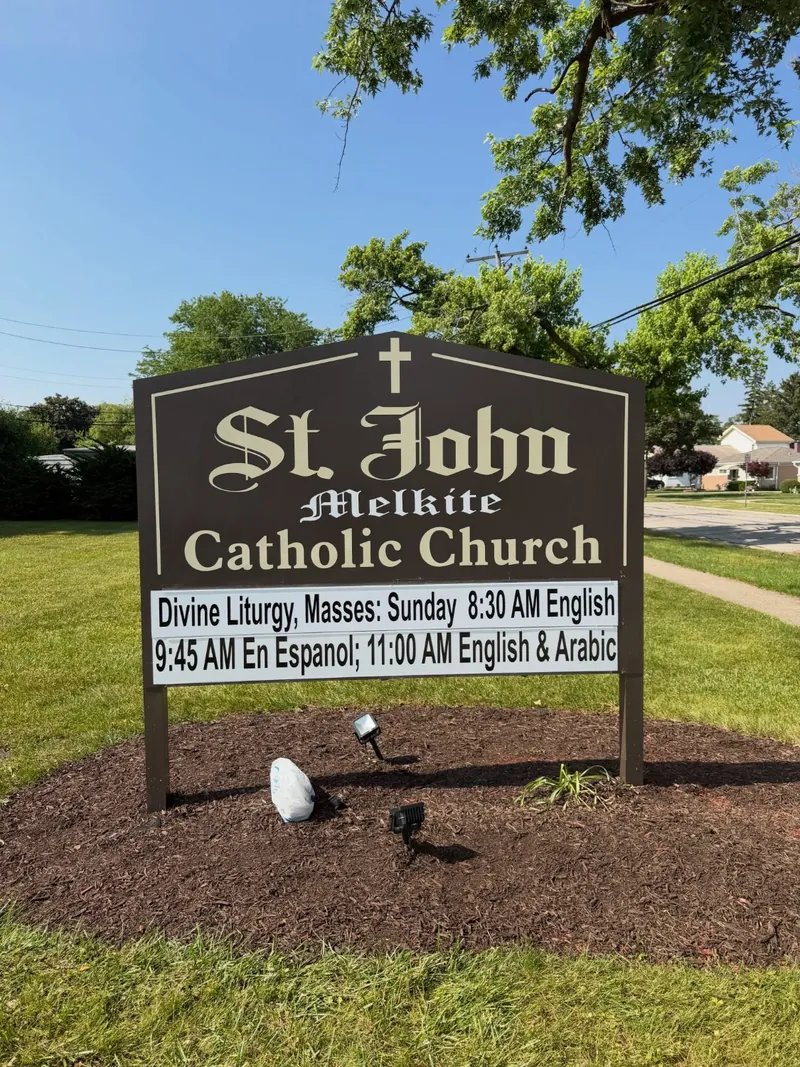 Brown monument sign with a cross for St. John Melkite Catholic Church, listing Sunday Divine Liturgy times