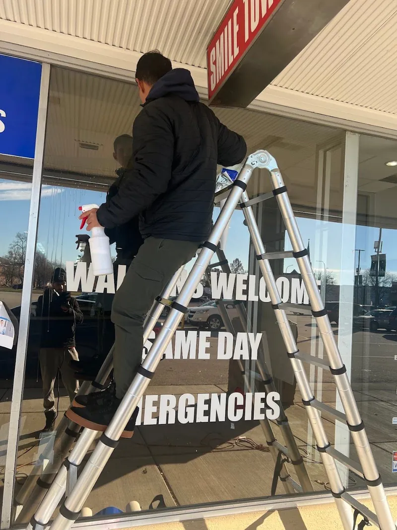Installer on a ladder applying white Walk-ins Welcome and Same Day Emergencies lettering to a glass storefront