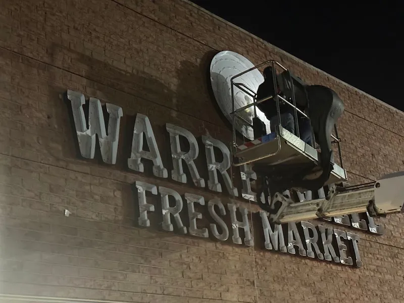 Lift raised against a brick façade at night while silver Warrenville Fresh Market letters are mounted