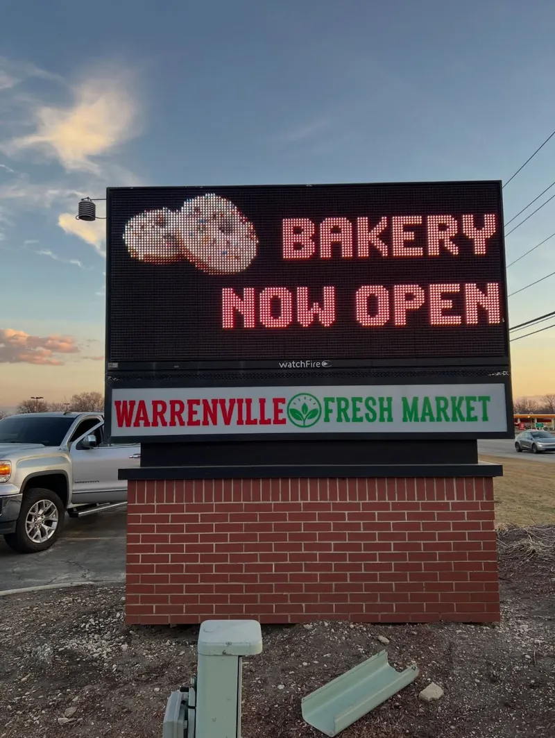 Pylon sign at dusk with an LED board reading Bakery Now Open above a Warrenville Fresh Market panel